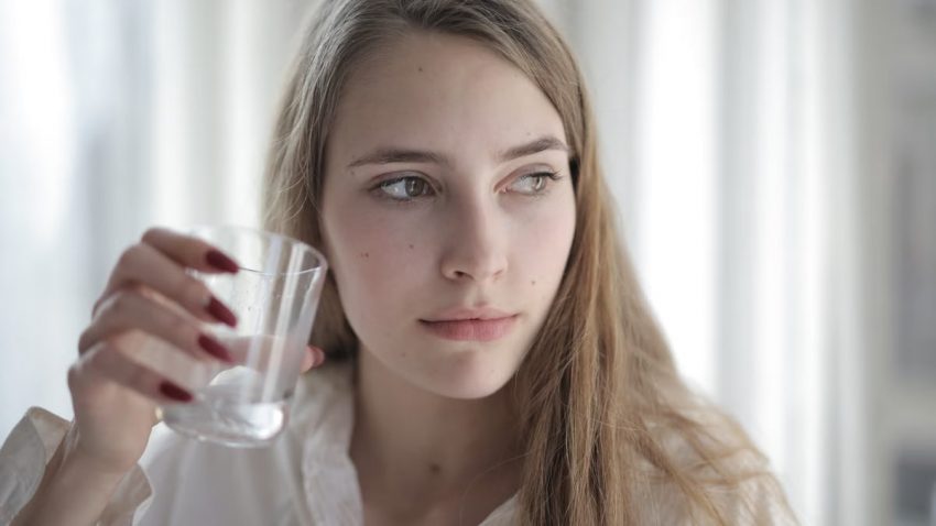 Young woman with long hair holding a glass of water, looking thoughtful.