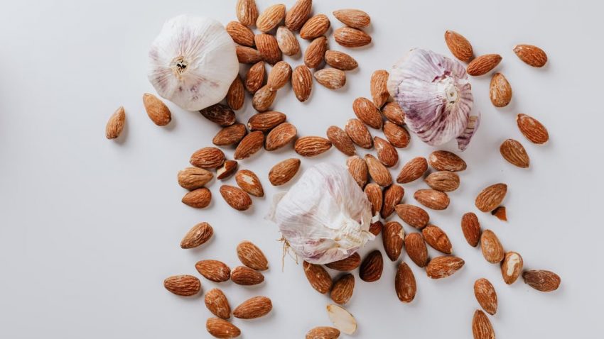 Top view of pile of raw partially peeled almonds and whole garlic bulbs with dry husk on white surface